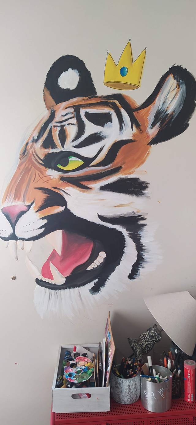 Wall mural of a roaring tiger head wearing a small gold crown, painted in orange, black and white above a desk of art supplies.