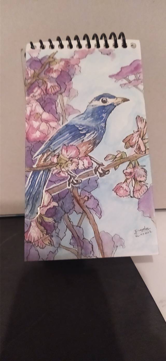 Watercolor of a blue bird perched among pink blossoms on a small spiral pad.