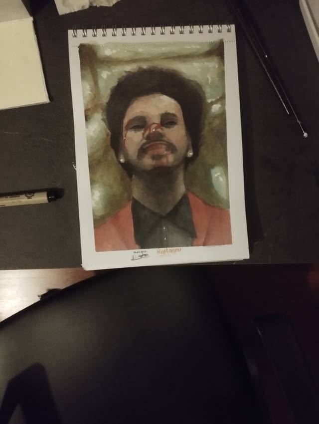 Watercolor portrait of a man with dark curly hair, a moustache and a red jacket on a spiral pad, photographed on a dark desk.