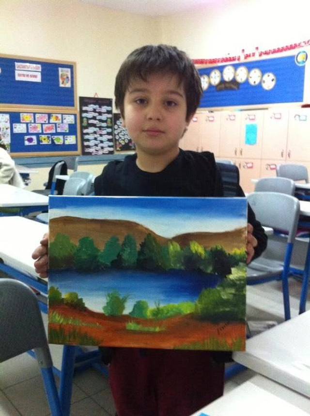 Photo of a young boy in a classroom holding up an acrylic landscape painting of green trees, a lake and a brown hillside.