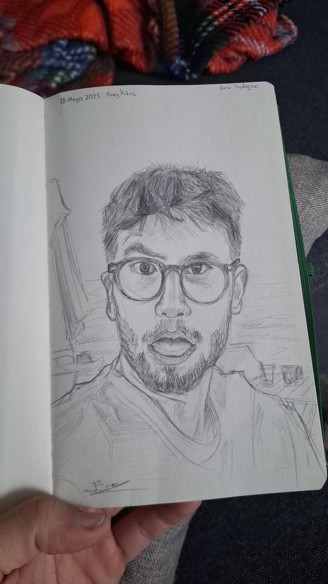 Pencil portrait of a bearded man with round glasses in a small sketchbook held open in one hand.